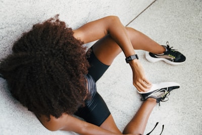 A person with curly hair is sitting on the ground with one leg extended and the other bent. They are wearing black athletic shorts and sneakers, and have a fitness tracker on their wrist.