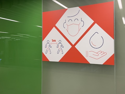 A sign with three sections uses simple line drawings to convey health safety measures. The first section shows two figures standing 1 meter apart, indicating social distancing. The second section features a face with a medical mask, and the third shows a hand catching a droplet, suggesting hygiene practices.