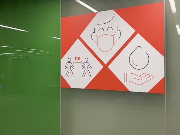 A sign with three sections uses simple line drawings to convey health safety measures. The first section shows two figures standing 1 meter apart, indicating social distancing. The second section features a face with a medical mask, and the third shows a hand catching a droplet, suggesting hygiene practices.