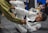 A person in a white martial arts gi is engaged in grappling, holding onto the leg of someone wearing a green gi. The scene takes place on a padded floor, with a few other people in the background observing.