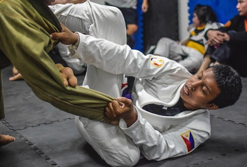 A person in a white martial arts gi is engaged in grappling, holding onto the leg of someone wearing a green gi. The scene takes place on a padded floor, with a few other people in the background observing.
