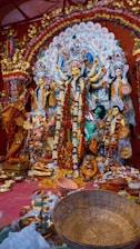 A vibrant and intricately decorated altar featuring a large idol of a goddess, possibly Durga, surrounded by smaller statues of other deities and figures. The scene is adorned with elaborate floral arrangements and offerings such as flowers, incense, and assorted items placed at the base. The backdrop is rich with detailed patterns and vivid colors.