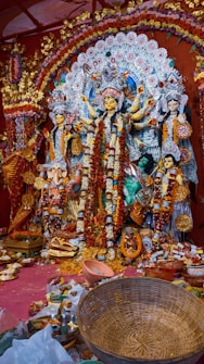A vibrant and intricately decorated altar featuring a large idol of a goddess, possibly Durga, surrounded by smaller statues of other deities and figures. The scene is adorned with elaborate floral arrangements and offerings such as flowers, incense, and assorted items placed at the base. The backdrop is rich with detailed patterns and vivid colors.