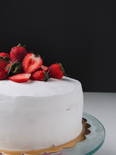 A round cake with smooth white frosting topped with fresh strawberries, some of which are halved. The cake is placed on a glass stand against a dark background.