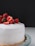 A round cake with smooth white frosting topped with fresh strawberries, some of which are halved. The cake is placed on a glass stand against a dark background.