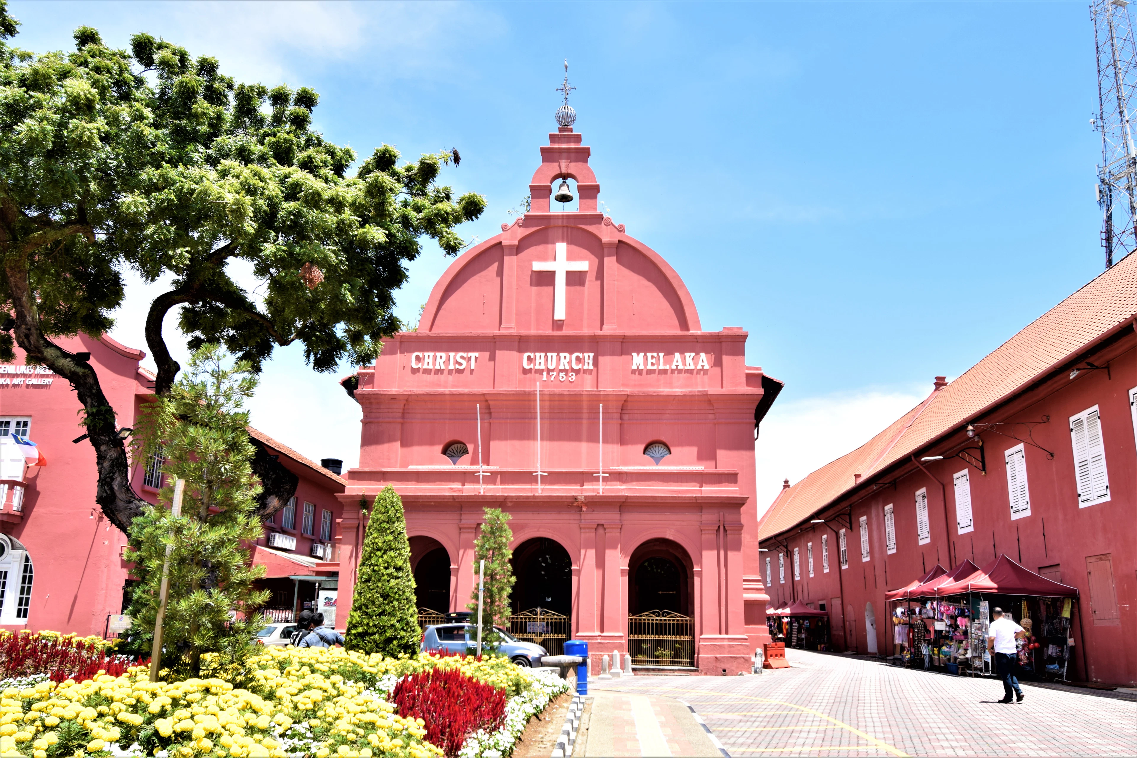The deep red Dutch colonial facade of Christ Church Melaka on Dutch Square in Malacca, Malaysia, its name painted beneath a white cross above beds of yellow and red flowers.