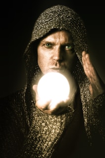 A person wearing a hooded garment is holding a glowing white orb in one hand, with a focused and intense expression. The surroundings are dark, enhancing the brightness of the orb.