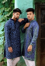 Two men wearing intricately patterned blue traditional outfits pose confidently against a textured green wall. They both have styled hair and are accessorized with rings. The scene includes lush green leaves trailing down the wall beside them.