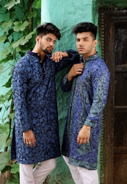Two men wearing intricately patterned blue traditional outfits pose confidently against a textured green wall. They both have styled hair and are accessorized with rings. The scene includes lush green leaves trailing down the wall beside them.