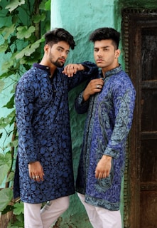 Two men wearing intricately patterned blue traditional outfits pose confidently against a textured green wall. They both have styled hair and are accessorized with rings. The scene includes lush green leaves trailing down the wall beside them.