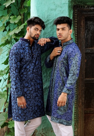 Two men wearing intricately patterned blue traditional outfits pose confidently against a textured green wall. They both have styled hair and are accessorized with rings. The scene includes lush green leaves trailing down the wall beside them.