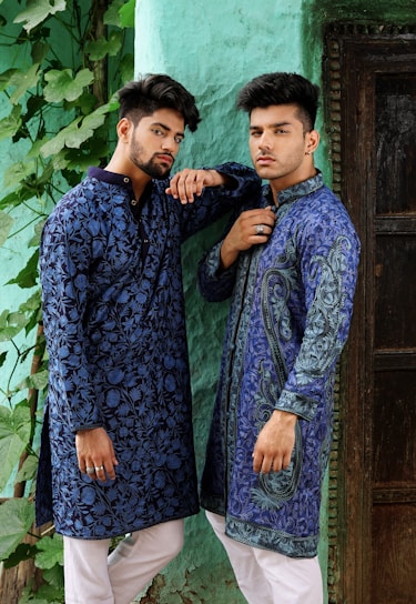 Two men wearing intricately patterned blue traditional outfits pose confidently against a textured green wall. They both have styled hair and are accessorized with rings. The scene includes lush green leaves trailing down the wall beside them.