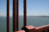 Several thick, rust-colored cables stretch vertically with a cityscape in the background across a body of water. A small sailboat can be seen on the water under a clear, bright blue sky.