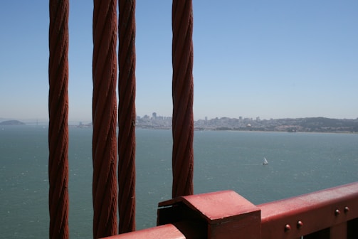 Several thick, rust-colored cables stretch vertically with a cityscape in the background across a body of water. A small sailboat can be seen on the water under a clear, bright blue sky.
