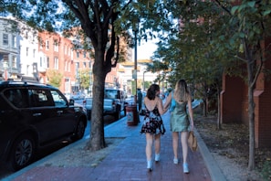 Two women in summer dresses walk arm-in-arm down a city sidewalk lined with trees and parked cars. The surrounding area includes buildings with restaurants and shops, creating a vibrant urban atmosphere.