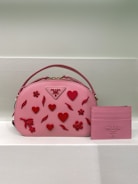 A pink handbag adorned with red heart and flower embellishments is paired with a matching pink cardholder. The brand logo is visible on both items.