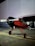 A red and black single-engine propeller airplane is parked on a concrete surface. It has a high wing design and tailwheel landing gear. The background consists of a large light-colored wall.