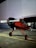 A red and black single-engine propeller airplane is parked on a concrete surface. It has a high wing design and tailwheel landing gear. The background consists of a large light-colored wall.