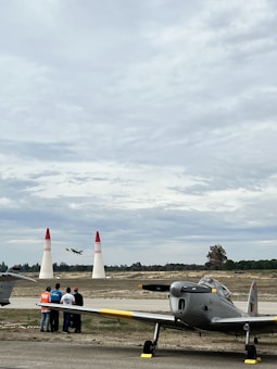 Several people stand near an aircraft on the tarmac, observing a small plane flying between two red and white pylons. The scene takes place under a cloudy sky, with trees visible in the distant background.