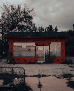 A small, abandoned building with boarded windows and a door, surrounded by overgrown vegetation. The building has red walls with black spots and is set against a cloudy, overcast sky. The ground in front is cracked and unkempt, with weeds growing through the pavement.