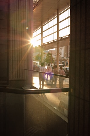 A warm, bright sun flares through the glass facade of a building, casting golden light on the interior and illuminating the silhouettes of people walking outside. The setting creates an interplay of shadows and reflections on the polished surfaces of the architecture.