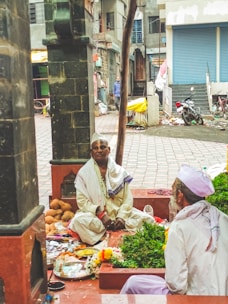Two individuals are sitting in a partially enclosed space, which appears to be a small temple or sacred area. One person is dressed in traditional white clothing and is seated on the ground with ritual items like coconuts, flowers, and plates around them. The other person, also in traditional attire, is sitting opposite, possibly engaged in a religious ritual or ceremony. The surroundings include a tiled floor, some plants, and a building in the background with a partially opened shutter.