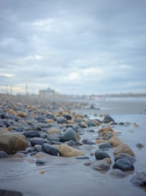 A rocky shoreline with numerous smooth, rounded stones scattered across the sand. The sky is overcast with thick gray clouds, creating a moody and serene atmosphere. The background fades into a distant blur, adding to the calm yet somber feel.