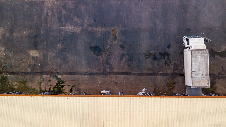 An aerial view of a white truck parked against a large dark concrete surface. The truck is located near the edge of a long, light-colored building roof, with industrial loading docks visible.