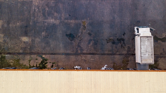 An aerial view of a white truck parked against a large dark concrete surface. The truck is located near the edge of a long, light-colored building roof, with industrial loading docks visible.