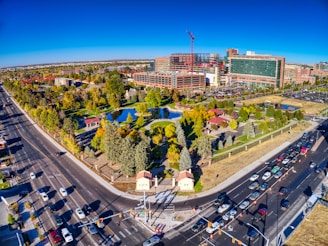 An aerial view of an urban area featuring a large park with lush green trees, a pond, and pathways. The park is surrounded by busy roads with visible traffic, leading to a modern hospital complex or office buildings under construction, identifiable by cranes and scaffolding.