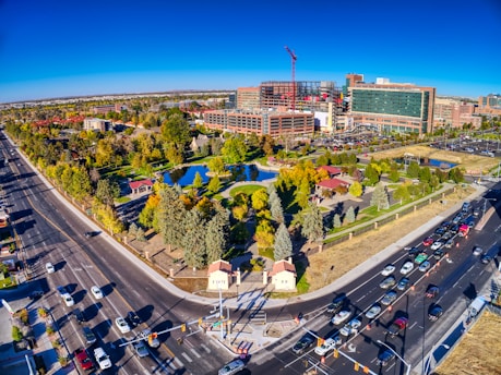 An aerial view of an urban area featuring a large park with lush green trees, a pond, and pathways. The park is surrounded by busy roads with visible traffic, leading to a modern hospital complex or office buildings under construction, identifiable by cranes and scaffolding.