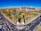 An aerial view of an urban area featuring a large park with lush green trees, a pond, and pathways. The park is surrounded by busy roads with visible traffic, leading to a modern hospital complex or office buildings under construction, identifiable by cranes and scaffolding.