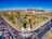 An aerial view of an urban area featuring a large park with lush green trees, a pond, and pathways. The park is surrounded by busy roads with visible traffic, leading to a modern hospital complex or office buildings under construction, identifiable by cranes and scaffolding.