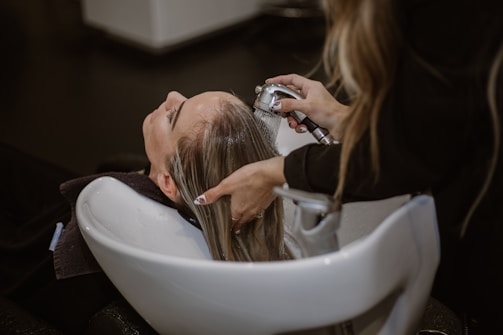 A person is having their hair washed in a salon setting. The stylist uses a handheld showerhead to rinse the hair over a white basin. The scene is calm and professionally arranged with focus on hair and hygiene.