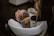 A person is having their hair washed in a salon setting. The stylist uses a handheld showerhead to rinse the hair over a white basin. The scene is calm and professionally arranged with focus on hair and hygiene.