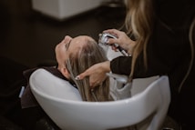 A person is having their hair washed in a salon setting. The stylist uses a handheld showerhead to rinse the hair over a white basin. The scene is calm and professionally arranged with focus on hair and hygiene.