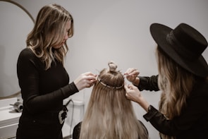 Two women are working on styling the hair of a third person whose back is facing the camera. One woman is using a tool, possibly to attach hair extensions or accessories, and the other is assisting. The setting appears to be a salon with neutral, calming colors.