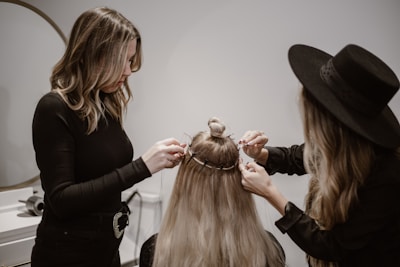 Two women are working on styling the hair of a third person whose back is facing the camera. One woman is using a tool, possibly to attach hair extensions or accessories, and the other is assisting. The setting appears to be a salon with neutral, calming colors.