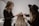 Two women are working on styling the hair of a third person whose back is facing the camera. One woman is using a tool, possibly to attach hair extensions or accessories, and the other is assisting. The setting appears to be a salon with neutral, calming colors.