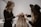 Two women are working on styling the hair of a third person whose back is facing the camera. One woman is using a tool, possibly to attach hair extensions or accessories, and the other is assisting. The setting appears to be a salon with neutral, calming colors.