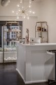 A beautifully organized shop interior featuring a modern chandelier with spherical lights hanging above a white reception desk. The desk has an Apple iMac computer on it, alongside some small decorative items such as flowers and cards. Behind the desk, there are shelves displaying various beauty and skincare products including lotions and bottles arranged neatly. The shelves have a vintage gold frame design and the room has a clean, minimalist aesthetic.