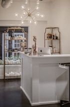 A beautifully organized shop interior featuring a modern chandelier with spherical lights hanging above a white reception desk. The desk has an Apple iMac computer on it, alongside some small decorative items such as flowers and cards. Behind the desk, there are shelves displaying various beauty and skincare products including lotions and bottles arranged neatly. The shelves have a vintage gold frame design and the room has a clean, minimalist aesthetic.