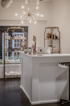 A beautifully organized shop interior featuring a modern chandelier with spherical lights hanging above a white reception desk. The desk has an Apple iMac computer on it, alongside some small decorative items such as flowers and cards. Behind the desk, there are shelves displaying various beauty and skincare products including lotions and bottles arranged neatly. The shelves have a vintage gold frame design and the room has a clean, minimalist aesthetic.