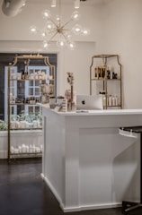 A beautifully organized shop interior featuring a modern chandelier with spherical lights hanging above a white reception desk. The desk has an Apple iMac computer on it, alongside some small decorative items such as flowers and cards. Behind the desk, there are shelves displaying various beauty and skincare products including lotions and bottles arranged neatly. The shelves have a vintage gold frame design and the room has a clean, minimalist aesthetic.