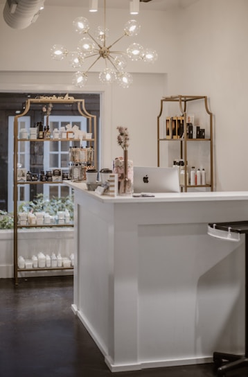 A beautifully organized shop interior featuring a modern chandelier with spherical lights hanging above a white reception desk. The desk has an Apple iMac computer on it, alongside some small decorative items such as flowers and cards. Behind the desk, there are shelves displaying various beauty and skincare products including lotions and bottles arranged neatly. The shelves have a vintage gold frame design and the room has a clean, minimalist aesthetic.