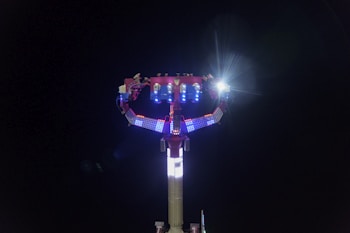 A brightly lit amusement ride with colorful LED lights against a dark night sky. The ride features a tall structure with arms extending outward and a bright light at the top.