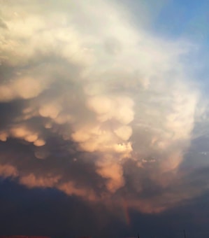Large, billowing clouds dominate the sky, featuring a mix of soft, rounded shapes with a gentle glow. The cloud formation displays various shades blending from light to darker hues, creating a dynamic and textured appearance.