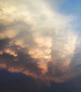 Large, billowing clouds dominate the sky, featuring a mix of soft, rounded shapes with a gentle glow. The cloud formation displays various shades blending from light to darker hues, creating a dynamic and textured appearance.