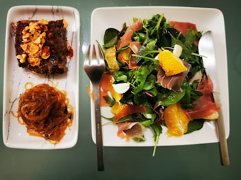 A visually appealing meal consisting of a rectangular plate with a tender beef slice topped with crushed nuts and a pile of seasoned noodles. Adjacent to it is a square plate featuring a fresh salad, adorned with leafy greens, slices of prosciutto, and segments of orange, accompanied by a fork and knife.
