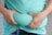 A person is holding their midsection, gripping the fabric of a turquoise shirt, emphasizing the belly area. The hands are positioned on both sides, highlighting body fat.